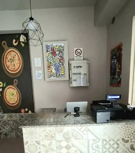 Il bancone del locale rivestito di maioliche, con la cassa, i quadri e il murale delle pizze alla parete